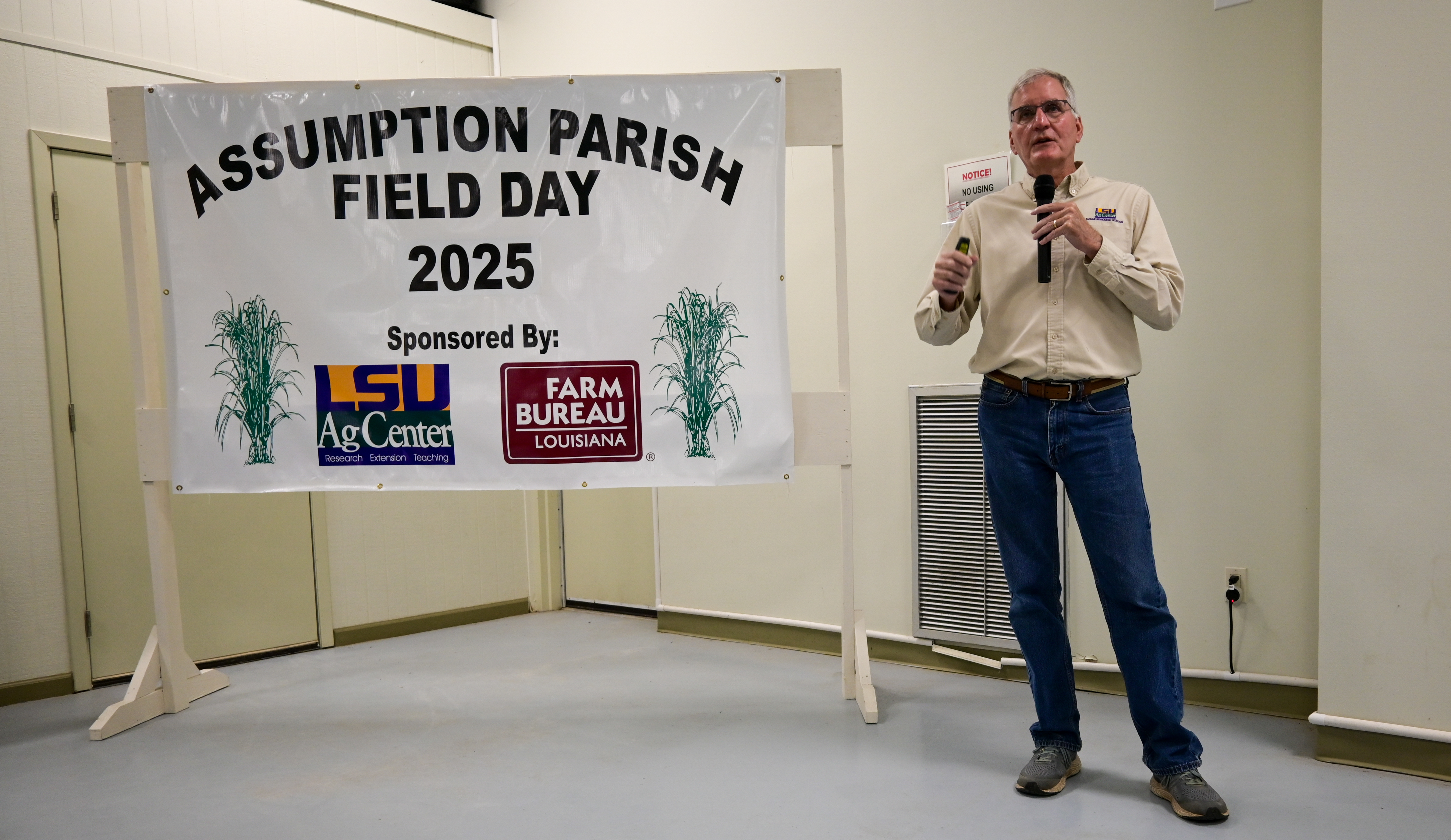 Man speaking while holding microphone and standing next to sign reading "Assumption Parish field day 2025".